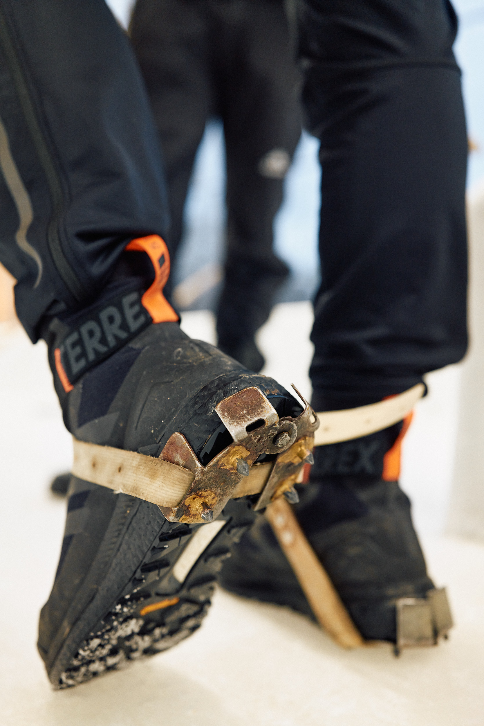 Eine Nahaufnahme von zwei Schuhen, an denen spezielle Spikes befestigt sind. Die Schuhe sind schwarz mit orangefarbenen Akzenten und werden durch Metallhalterungen an den Spikes fixiert. Der Boden scheint eisbedeckt zu sein. 