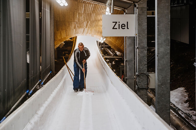 Ein Mann steht auf einer Eisbahn und reinigt die Bahn mit einem Werkzeug. Über ihm hängt ein Schild mit der Aufschrift „Ziel“. Die Eisbahn ist von einer hölzernen Überdachung umgeben und gut beleuchtet.