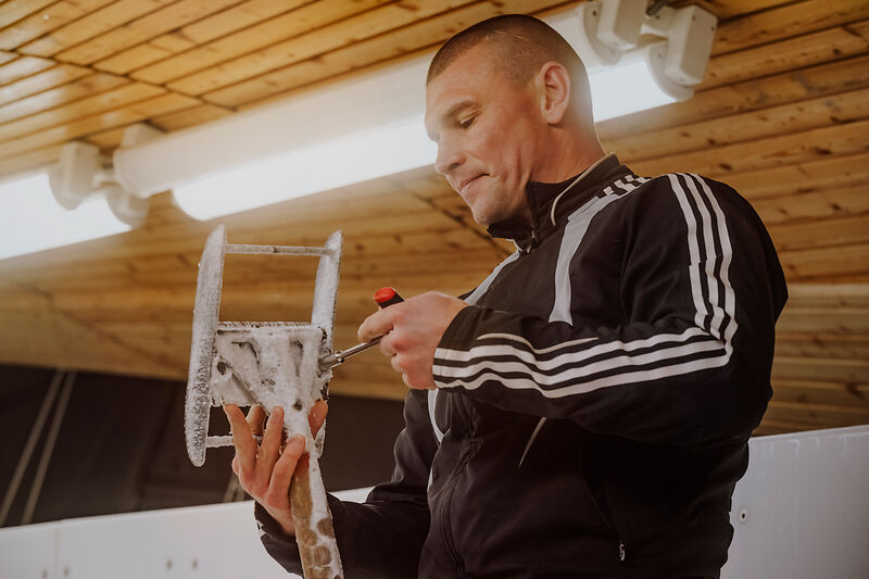 Ein Mann in einer schwarzen Trainingsjacke hält ein vereistes Metallwerkzeug in den Händen. Er verwendet einen Schraubendreher, um an dem vereisten Gerät zu arbeiten. Im Hintergrund ist eine Holzdecke mit Beleuchtung zu sehen. 