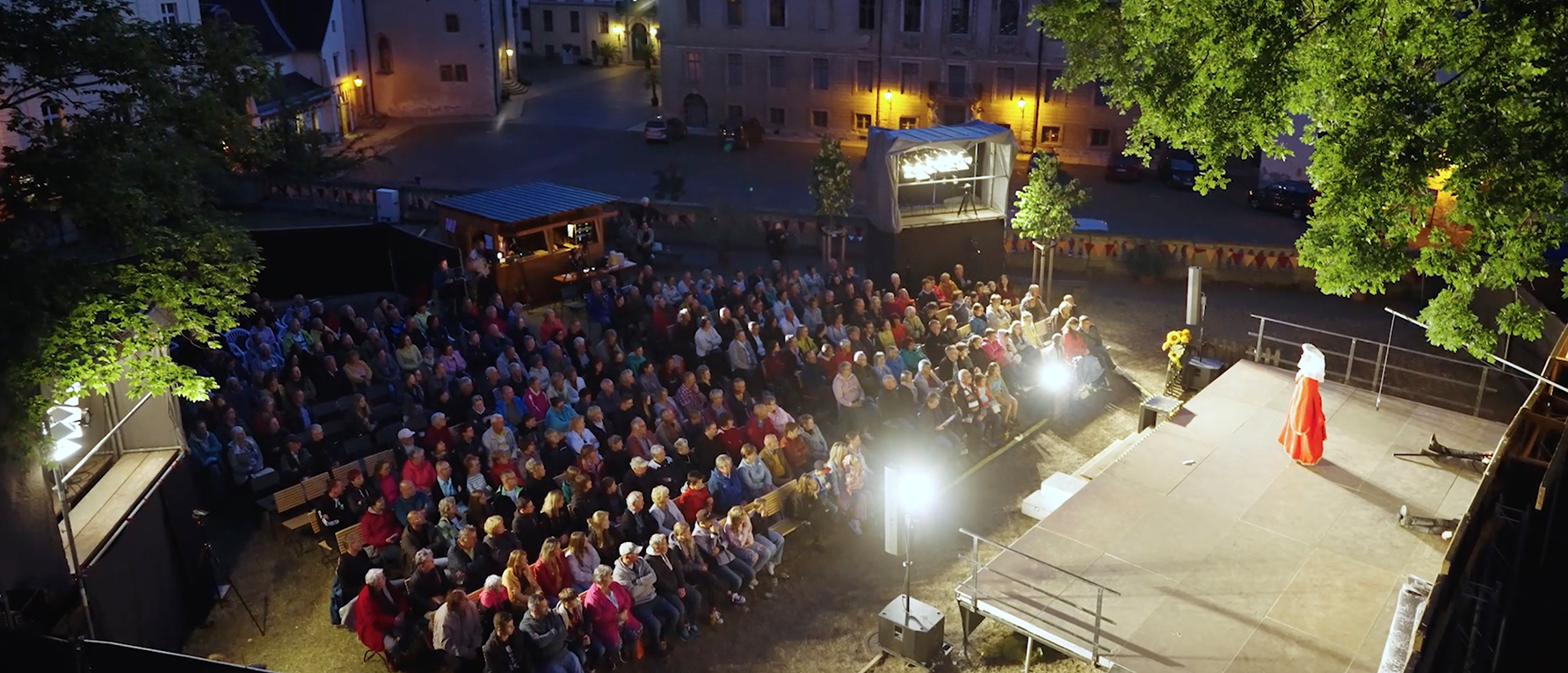 Ein Blick wird von oben auf das Theaterstück der Altenburger Prinzenraub-Festspiele geworfen. Die sitzenden Zuschauer schauen bei abendlichem Licht auf die angestrahlte Bühne, auf der eine Schauspielerin im roten Kostüm und weißem Hut steht. Im Hintergrund erkennt man die Kulisse des Schlosses von Altenburg.