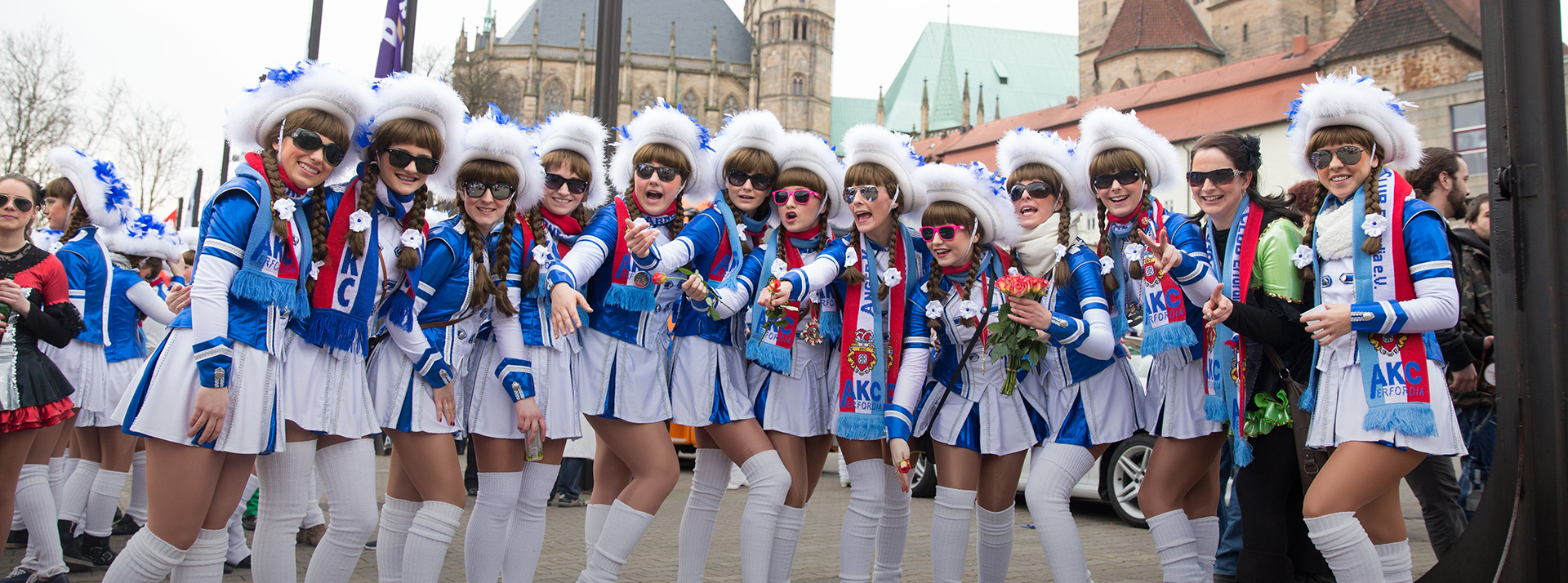 Eine Gruppe von Karnevalmädchen posiert auf einem Umzug im Jahr 2014 für die Kamera. Die Mädchen sind vom Anger Karneval Club Erfordia e.V. (AKC) und tragen blau-weiße Kostüme, weiße Stulpen und weiß-blaue Hüte. Dazu trägt die Gruppe Sonnenbrillen.