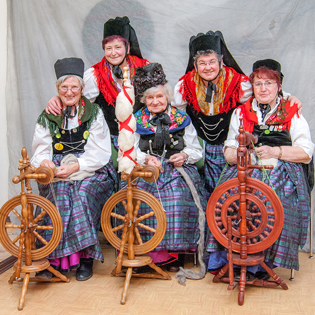 Ein Gruppenbild von fünf älteren Frauen in der Friemar Tracht. Die vorderen drei Frauen sitzen jeweils an einem Spinnrad. Die anderen beiden haben sich für das Bild vorgelehnt. Die Frauen tragen ein dunkles ärmellose Kleid mit einer lila-grün-karierten Schürze über einer weißen Bluse. Dazu haben die Frauen bunte Dreieckstücher über die Schultern geworfen und tragen schwarze Kappen.