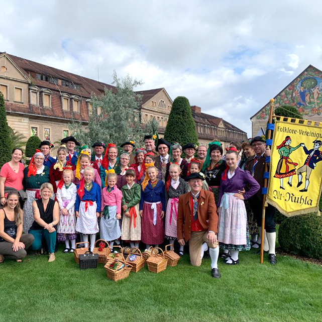 Ein Gruppenbild des Trachtenvereins Alt-Ruhla im Grünen. Mehrere Leute von jung bis alt posieren in ihrer bunten Tracht. Rechts hält ein Mann eine gelbe Fahne mit der Aufschrift „Folklore-Vereinigung Alt-Ruhla“ und zwei gemalten Personen in Tracht.