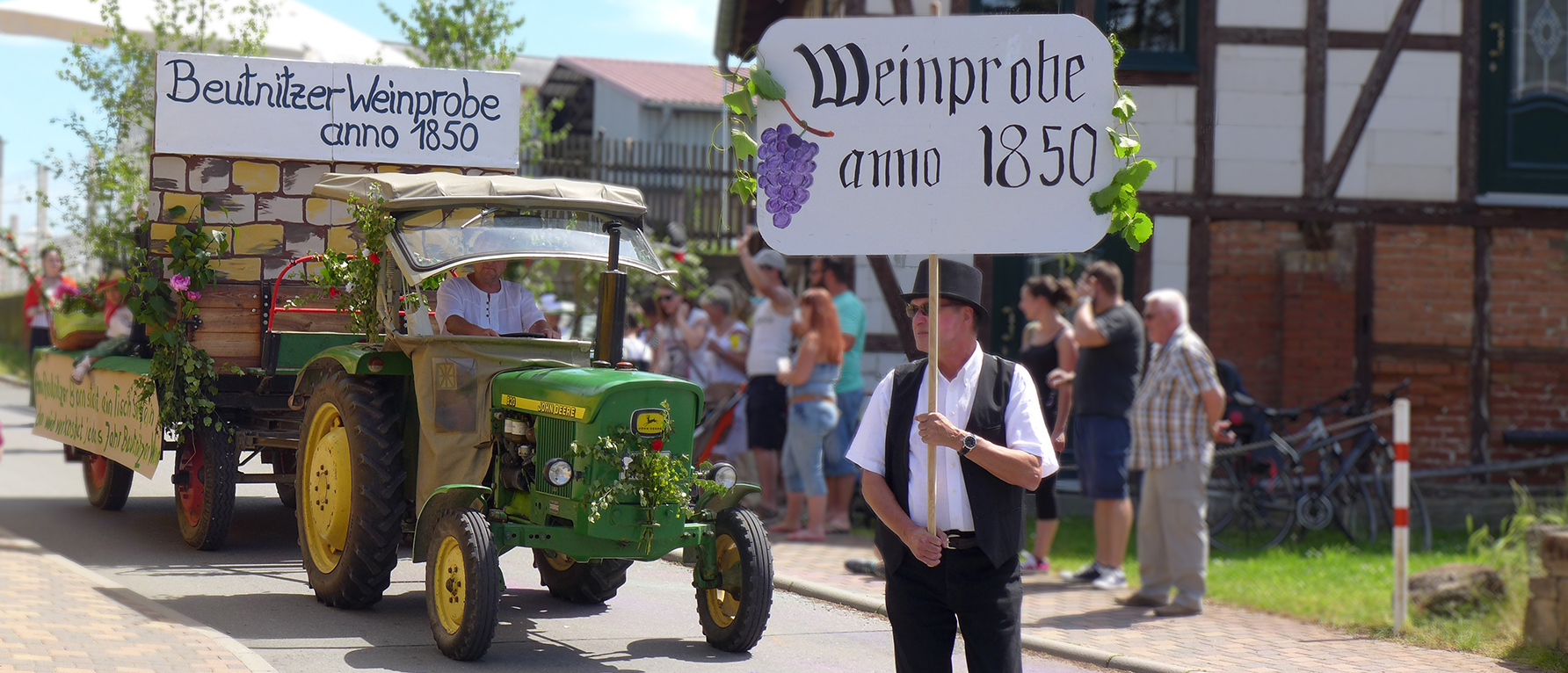 Ein Mann steht auf einer Dorfstraße und hält ein Schild hoch mit der Aufschrift: „Weinprobe anno 1850“. Im Hintergrund stehen Schaulustige, während ein Traktor mit einem geschmückten Festanhänger vorbeifährt. Auf einem Schild am Anhänger steht „Beutnitzer Weinprobe anno 1850“. 