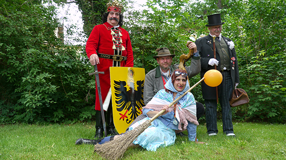 Die vier Figuren des Rolandsfest posieren für ein Foto im Gras. Vorne liegt eine Frau im hellblauen Hexenkostüm mit Hakennase und Besen, die die Figur der Brockenhexe verkörpert. Links hinter ihr steht die Figur des Rolands, ein korpulenter Mann in langem rotem Mantel, Krone und markantem Bart. Er hält in der rechten Hand ein Schwert und in der linken Hand ein gelbes Schild mit Wappen. Direkt hinter der Brockenhexe hockt ein Mann im Jägerkostüm samt grauer Jacke und Jägerhut. Er verkörpert die Figur des Ebersberger. Rechts neben ihm steht ein älterer Mann, der die Figur des Professor Zwanziger darstellt. Er ist in einer schwarz-grau gestreifter Hose, dunkelgrüner Weste und einem schwarzem Wrack, mit Ansteckern gekleidet. Er trägt weiße Handschuhe, einen schwarzen Zylinder und eine runde Brille. In seiner linken Hand liegt eine Ledertasche und in seiner rechten ein edler, hölzerner Gehstock.