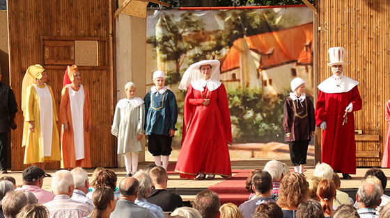  Die Theaterdarsteller der Altenburger Prinzenraub-Festspiele begeistern mit prachtvollen Kostümen auf der Bühne. Sie stehen aufgereiht nebeneinander. In der Mitte steht eine Frau in roter Robe und weißem Hut nebst zwei Jugendlichen in blauen und grauem Kleid und weißen Mützen. Die Zuschauer schauen sitzend gebannt auf die Freilichtbühne.