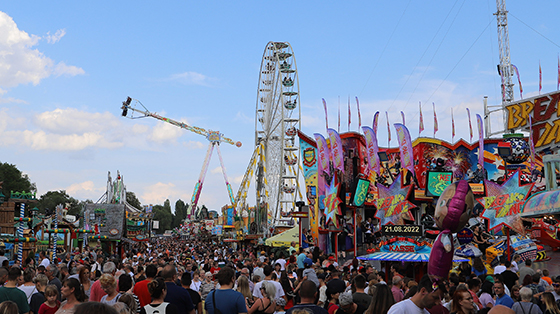 Eine Menschenmenge schlendert über das Rudolstädter Vogelschießen ähnliches eines Doms oder einer Kirmes. An der Seite stehen bunte Geschäfte mit Fahnen und im Hintergrund lässt sich ein Riesenrads und diverse bunte Fahrgeschäfte erkennen.