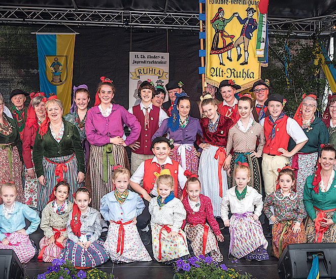 Ein Gruppenbild der Kirmesgesellschaft von Ruhla auf einer Bühne in bunten, individuellen Trachten. Im Hintergrund sieht man ein gelbes Banner auf dem zwei Figuren in Tracht gekleidet tanzen. Es trägt den Titel „Alt-Ruhla“ - in altertümlicher Schrift geschrieben.