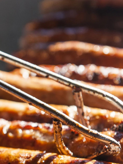 Mehrere Thüringer Rostbratwürste liegen auf einem Grill im Sonnenschein und werden mit einer Zange gewendet