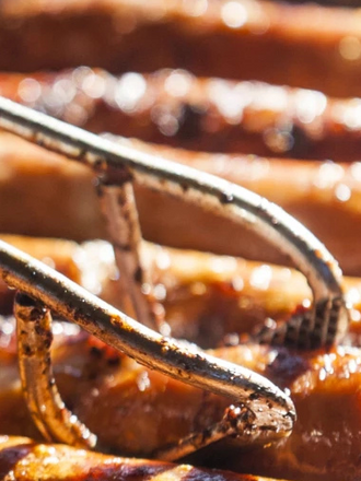 Mehrere Thüringer Rostbratwürste liegen auf einem Grill im Sonnenschein und werden mit einer Zange gewendet
