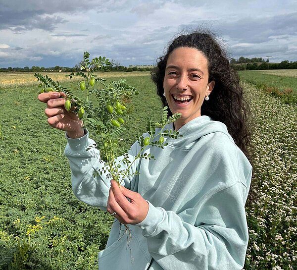 Ivana Fontanini, Gründerin von TAIAO, lacht vor einem Kichererbsen-Feld mit Kichererbsenpflanze in der Hand