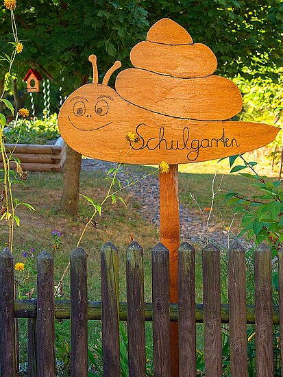 Am Holzlattenzaun eines Gartens mit Bäumen, Vogelhaus, langstieligen gelben Blumen und Holzeingefasstem Hochbeet mit Schild "Schulgarten" in Schneckensilhouette