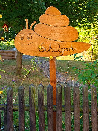 Am Holzlattenzaun eines Gartens mit Bäumen, Vogelhaus, langstieligen gelben Blumen und Holzeingefasstem Hochbeet mit Schild "Schulgarten" in Schneckensilhouette