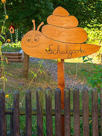 Am Holzlattenzaun eines Gartens mit Bäumen, Vogelhaus, langstieligen gelben Blumen und Holzeingefasstem Hochbeet mit Schild "Schulgarten" in Schneckensilhouette