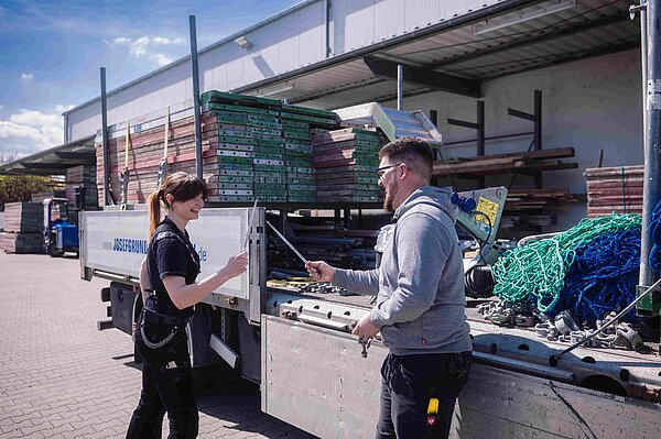 Ein Gerüstbauer und eine Gerüstbauerin stehen vor einem Sattelschlepper und liefern sich einen Schwertkampf mit zwei Bolzen