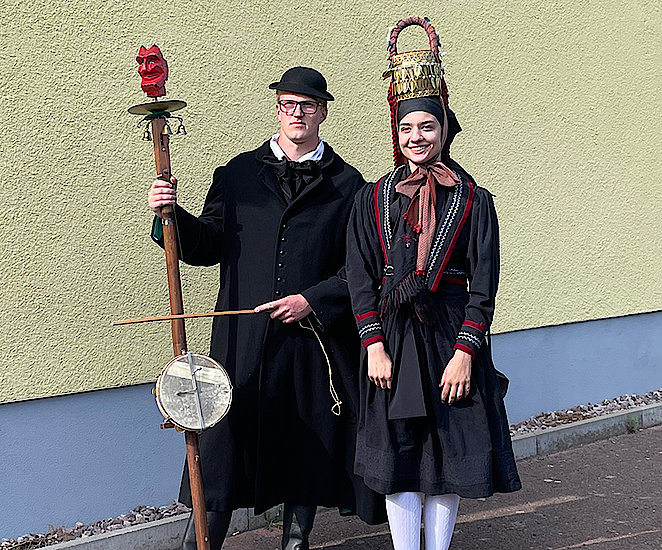 Zwei junge Erwachsene posieren vor einer Wand in Altenburger Tracht. Er steht links und trägt einen langen schwarzen Mantel, hohe schwarze Lederstiefel und einen schwarzen Hut mit kurzer Krempe. Sie trägt zu weißen Strümpfen ein schwarzes, hochgeschlossenes Kleid mit rot-weißem Rand und blassroter Schleife um den Hals. Auf ihrem Kopf trägt die Frau die markante Kopfbedeckung der Tracht: eine hohe Krone mit kleinen Blättern aus vergoldetem Silber dran mit einer Art Griff darüber. Diese Brautkorne wird goldenes Hormt genannt.