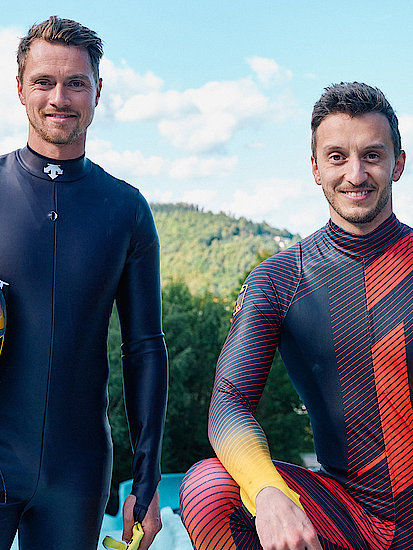 Johannes Ludwig und Sascha Benecken stehen nebeneinander und lächeln in die Kamera. Sie tragen ihre Rennrodelanzüge und tragen ihren Helm unter dem Arm. Im Hintergrund sieht man die Rennrodelbahn und Ilmenau.