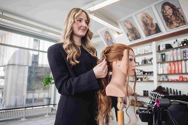 Eine blonde junge Frau im schwarzen Blazer übt an einem Puppenkopf Hochsteckfrisuren
