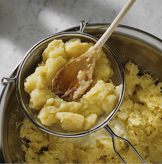 In einer silbernen Schale liegt ausgewrungener und roher Kartoffelrieb mit Butter. Über der Schale liegt ein Sieb, in dem gekochte Kartoffeln gestapelt sind, die mithilfe eines Kochlöffels aus Holz durch das Sieb gedrückt werden.