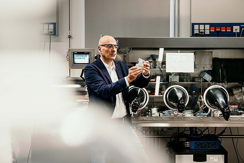 Ein Mann mit Schutzbrille steht in einem Raum mit verschiedenen technischen Apparaturen und hält einen Gegenstand aus Metall in den Händen.