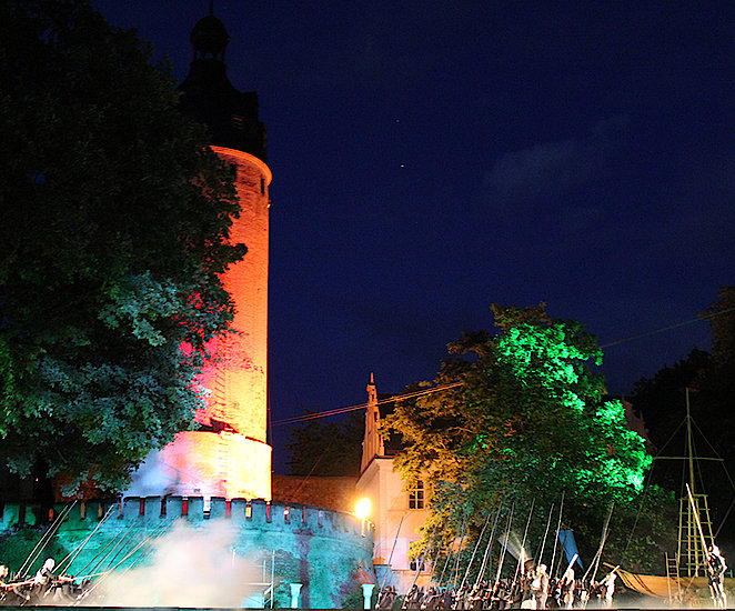 Das Schloss Altenburg wird am Abend der Aufführung mit bunten Lichtern angestrahlt. Im Vordergrund wird das Theaterstück aufgeführt.