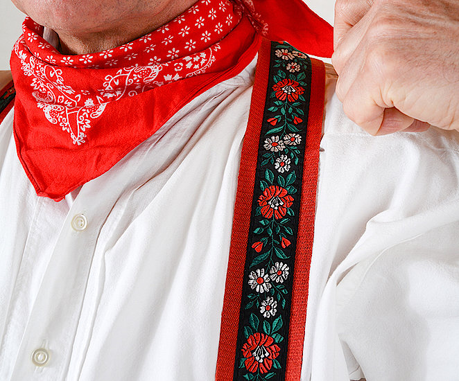 Ein Blick auf die Details der Hosenträger der Männertracht von Stepfershausen. Über einem weißen Hemd sieht man die schwarzen Hosenträger mit rotem Rand und rot-weißen Blumenstickereien. Der Mann auf dem Foto trägt dazu ein rotes Bandana um den Hals geknotet.