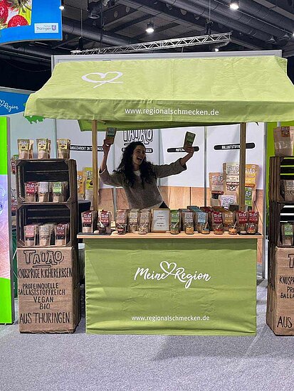Messestand von TAIAO in Marktstand-Optik mit grüner Zeltdecke, aufgereihten Produkten und lachender Start-up-Unternehmerin Ivana Fontanini