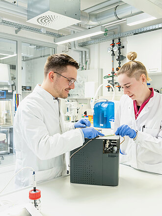 Zwei Personen in weißen Kitteln und mit blauen Handschuhen arbeiten in einem Labor gemeinsam an einem Gerät. Im Hintergrund befinden sich verschiedene technische Geräte und Apparaturen.
