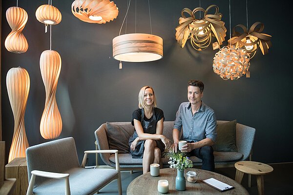 Franziska und Stefan Weiland sitzen auf einem Sofa vor grauer Wand und unter Lampenschirmen in nordischem Design