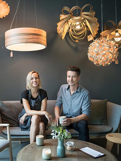 Franziska und Stefan Weiland sitzen auf einem Sofa vor grauer Wand und unter Lampenschirmen in nordischem Design