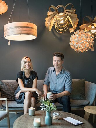 Franziska und Stefan Weiland sitzen auf einem Sofa vor grauer Wand und unter Lampenschirmen in nordischem Design