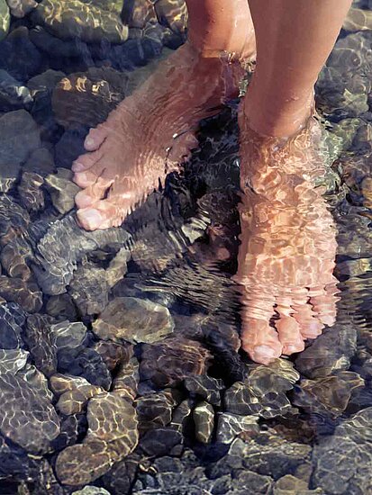Füße einer Frau bis zum Knöchel in Wasser, auf dessen Grund Kieselsteine liegen
