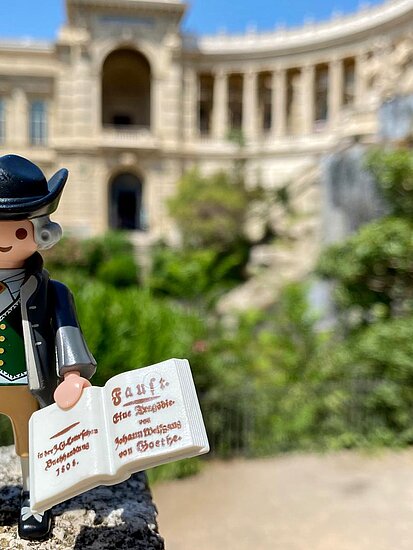 Eine PLAYMOBIL-Figur von Goethe mit einem schwarzen Hut und einer Feder in der einen sowie einem aufgeschlagenen Buch in der anderen Hand steht vor einem großen Gebäude, mit mehreren Fenstern und Säulen. Vor dem Gebäude ist eine Art Parkanlage zu erkennen. 