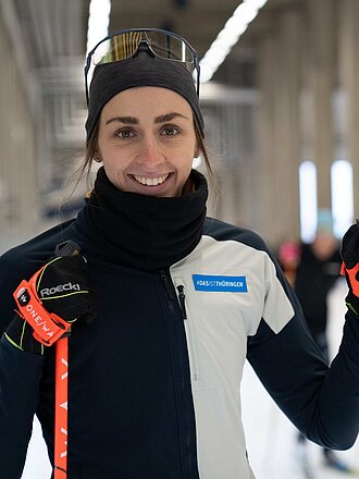 Vanessa Voigt in Trainingsoutfit und mit Ski-Stock schaut lächelnd in die Kamera in der Oberhofer Skihalle