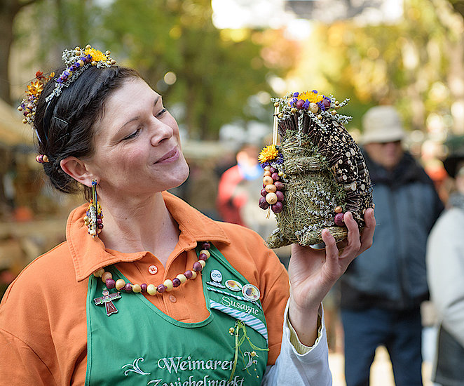 Eine Frau hält eine Igelfigur aus Holz und zusammengebundenen Heu in der Hand, die mit Zwiebeln verziert ist. Die Frau trägt ein oranges T-Shirt und eine grüne Schürze mit der Aufschrift „Weimarer Zwiebelmarkt“ mit ihrem Namen „Susanne“. Ihre Kette, Ohrringe und Haarschmuck bestehen aus kleinen Zwiebeln.