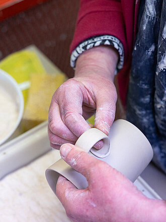 Zwei Händen befestigen einen gebogenen Henkel aus Porzellanmasse an einen Tassenrohling