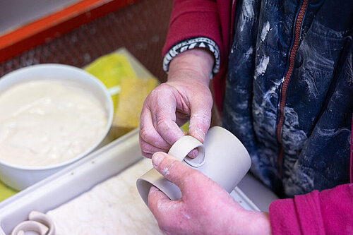 Zwei Händen befestigen einen gebogenen Henkel aus Porzellanmasse an einen Tassenrohling