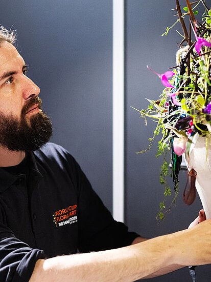 Florist mit Vollbart und dunklem Sweater arrangiert ein hängendes weißes Blumengefäß in einer Ausstellung vor dunklen Wänden