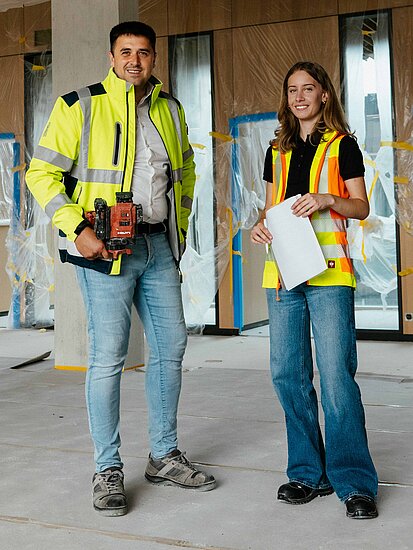 Ein Mann und eine Frau mit Warnjacke, Papier und Werkzeug in der Hand blicken lächelnd in die Kamera auf einer Baustelle im Innenausbau