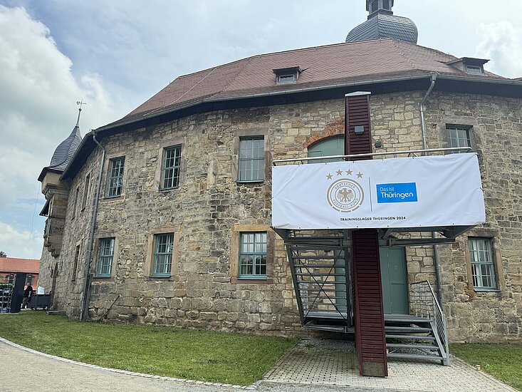 Ein zweigeschossiges Gebäude mit Steinfassade. Am Rand befindet sich eine Treppe, an deren Geländer ein weißes Banner angebracht ist. Auf diesem sind ein rundes, goldenes Logo sowie ein rechteckiges, blaues Logo zu sehen.