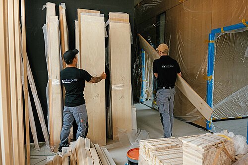Auf einer Innenraum-Baustelle tragen zwei Männer in Arbeitskleidung mit der Aufschrift "ruben peter" lange Holzplatten von einer Lagerstätte weg