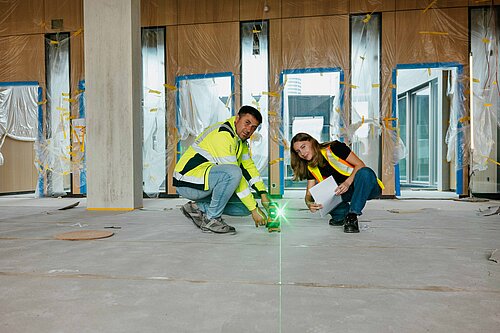 Auf einer Innenraum-Baustelle tragen zwei Männer in Arbeitskleidung mit der Aufschrift "ruben peter" lange Holzplatten von einer Lagerstätte weg