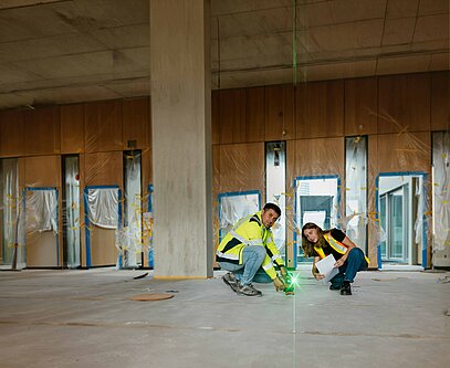 Auf einer Innenraum-Baustelle tragen zwei Männer in Arbeitskleidung mit der Aufschrift "ruben peter" lange Holzplatten von einer Lagerstätte weg