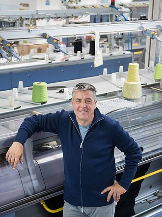 Geschäftsführer von Strick Zella, Dr. Gottfried Betz, steht in der Manufaktur vor den Strickmaschinen und blickt nach oben in die Kamera