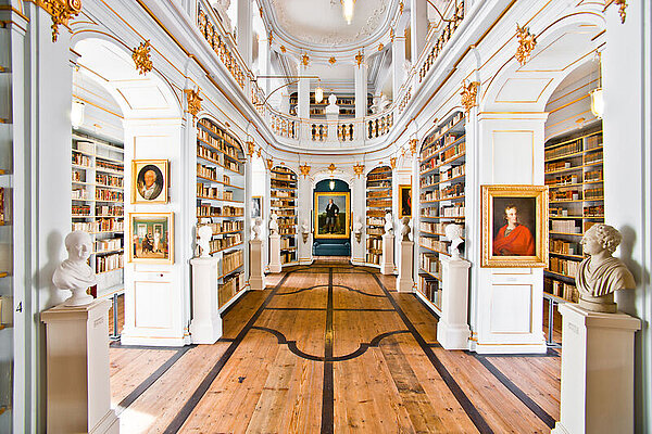 Blick in den zweigeschossigen weiß-vergoldeten Rokokosaal der Anna Amalia Bibliothek mit Parkett, Bücherregalen und Gemälden