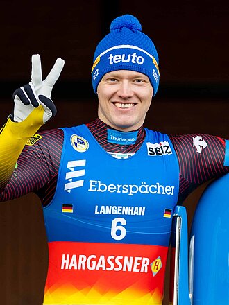 Max Langenhan mit Arm auf Rodelbrett und in Sportkleidung lächelt in die Kamera und zeigt zwei Finger als V