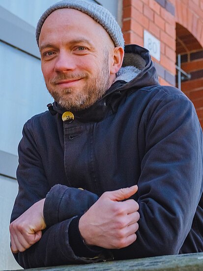 Dirk Rauscher trägt eine Wollmütze und schaut lächelnd vor einem Fachwerkhaus in die Kamera