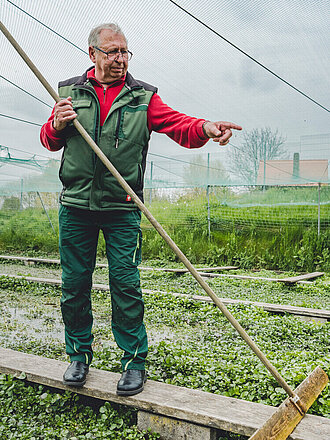 Der Oberkörper eines älteren Mannes in grüner Arbeitskleidung. Er hält ein langes Werkzeug in einer Hand und zeigt mit der anderen in die Ferne. Der Mann befindet sich unter einem Netz in einem landwirtschaftlichen Anbaugebiet und trägt eine Brille.