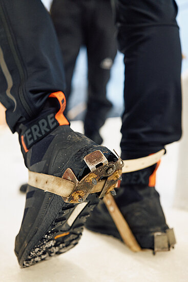 Eine Nahaufnahme von zwei Schuhen, an denen spezielle Spikes befestigt sind. Die Schuhe sind schwarz mit orangefarbenen Akzenten und werden durch Metallhalterungen an den Spikes fixiert. Der Boden scheint eisbedeckt zu sein. 