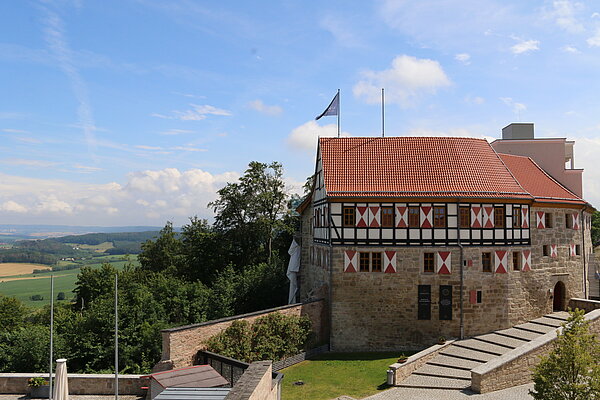 Burgpanorama mit Vorplatz und einem Haus aus hellem Stein und Fachwerk mit rot-weißen Fensterläden